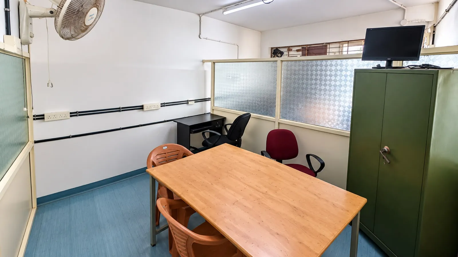 Cabin 3 at TBI MESCE: a wooden table with plastic and office chairs, a computer desk, frosted glass partitions and a green steel cupboard with a monitor on top