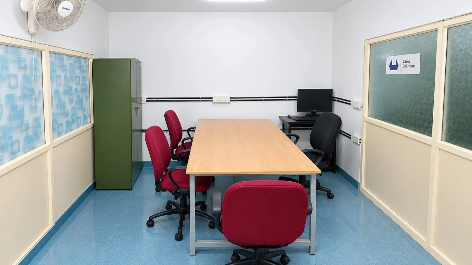 Cabin 5 at TBI MESCE: a long wooden table with red office chairs, a computer desk with a monitor in the corner, a green steel cupboard and a Zaina Solutions sign on the frosted glass partition